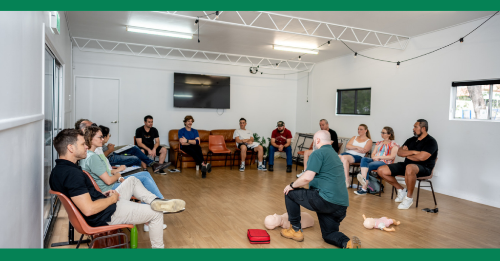 Another first aid training session with a different setting. Participants are seated in a semi-circle around an instructor demonstrating CPR on manikins. The room has large windows, and participants have various training materials around them.
