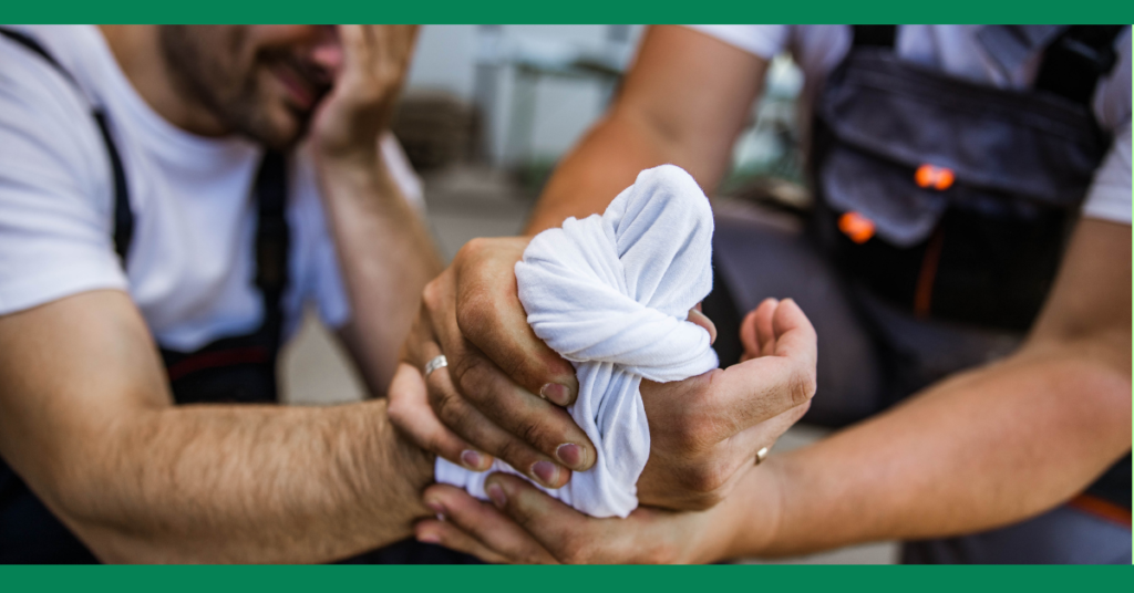 A close-up of two people applying a makeshift bandage to an injured hand using a piece of cloth. One person is holding the bandaged hand while the other assists.
