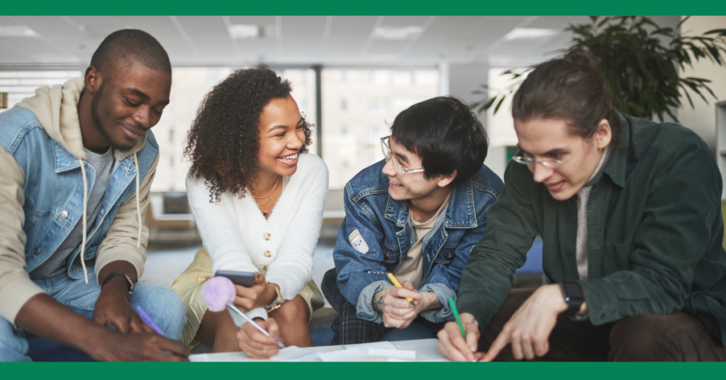 A group of young adults sitting together at a table, smiling and engaging in a collaborative activity. They appear to be working on something with notebooks and pens.
