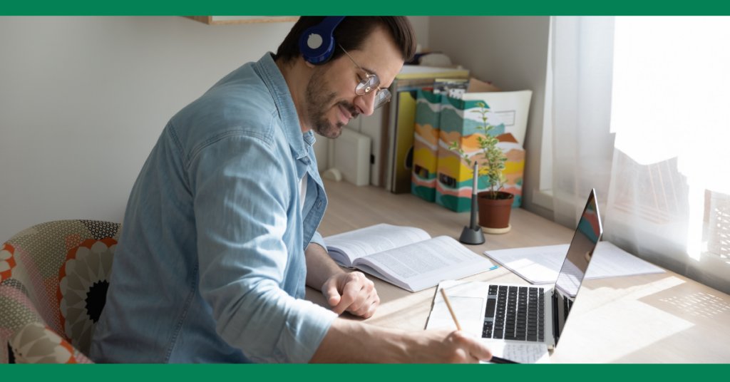 A man wearing headphones and glasses is studying at a desk with a laptop and books. He is writing notes while looking at the laptop screen.
