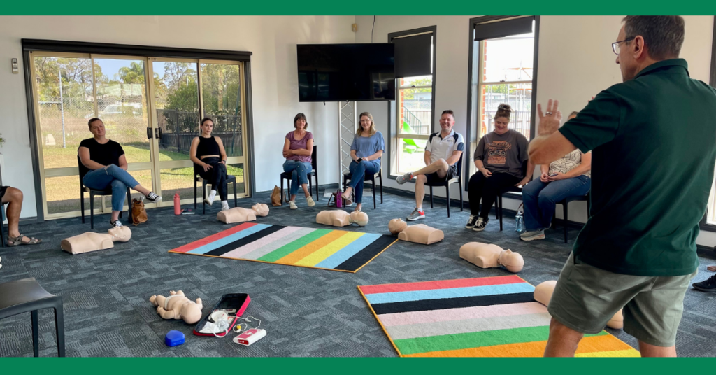 Another first aid training session similar to Image 3, but with a different setting. Participants are seated in a semi-circle around an instructor demonstrating CPR on manikins. The room has large windows, and participants have various training materials around them.
