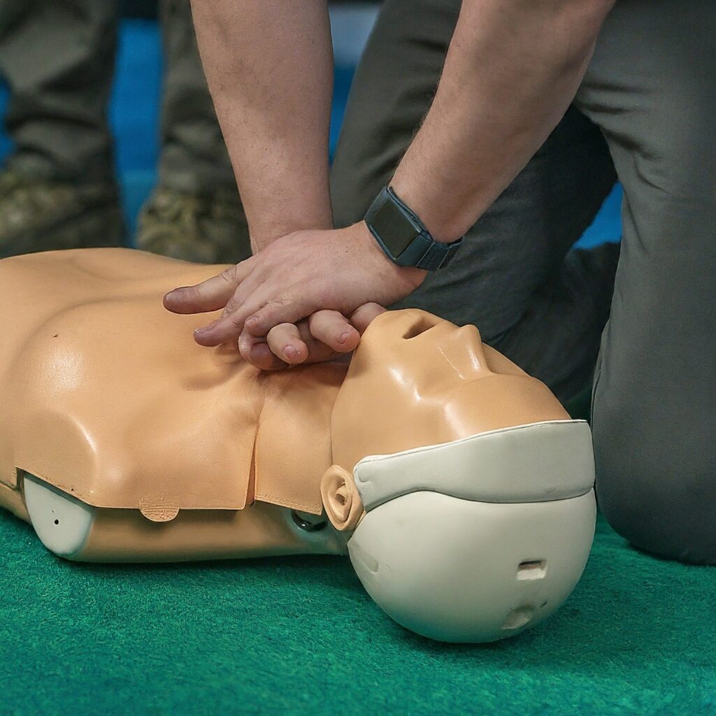 A person performing CPR chest compressions on a practice mannequin. The mannequin has a tan body and head, with a white face mask. The person is wearing a smartwatch on their wrist, and the background features a green surface with another person standing in the background.






