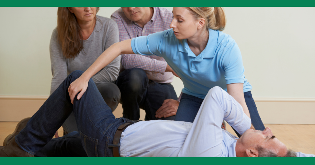 A woman wearing a light blue polo shirt demonstrates how to place a man in the recovery position. She is kneeling beside him, carefully positioning his arm and leg. Three other adults are observing closely, indicating a first aid training session. The background features a plain indoor setting with a light-colored wall and wooden floor.






