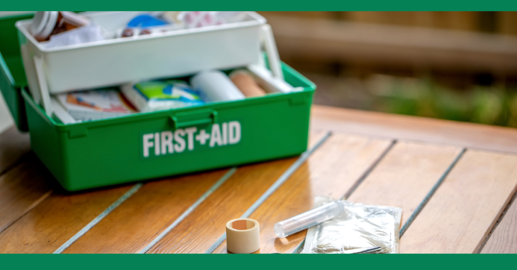 A close-up photograph of an open green first aid kit placed on a wooden table. The kit is filled with various medical supplies, such as bandages, antiseptics, and other first aid essentials. In the foreground, a few items, including a roll of adhesive tape, a syringe, and a packaged item, are spread out on the table. The background is slightly blurred, indicating an outdoor setting, possibly a backyard or garden. The image conveys readiness and the importance of having a well-stocked first aid kit available for emergencies.







