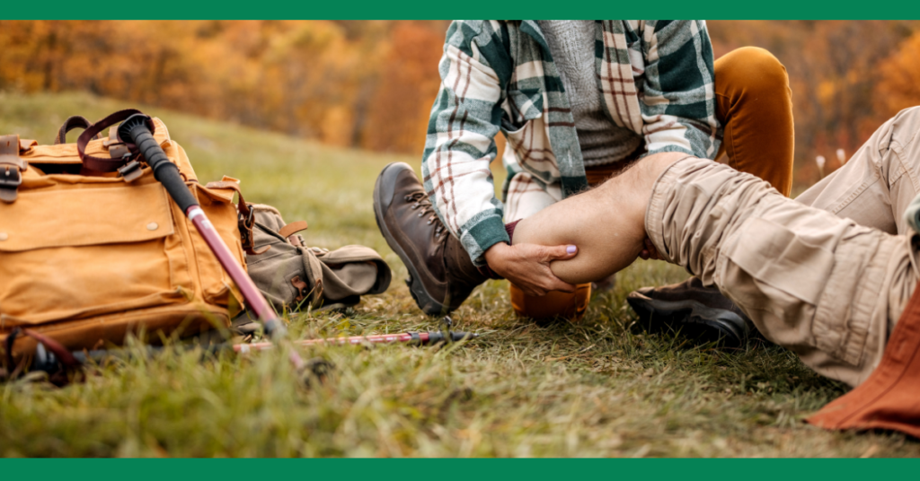  An outdoor scene where a person is receiving first aid for a leg injury while hiking, with a backpack and trekking poles visible in the background.