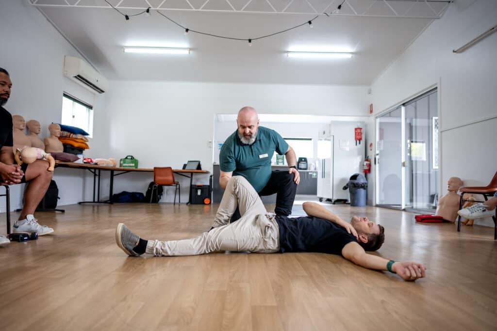 Step 2 of the recovery position: A first aid instructor assists a person lying on the floor, starting to position them into the recovery position, with another person observing in the background.