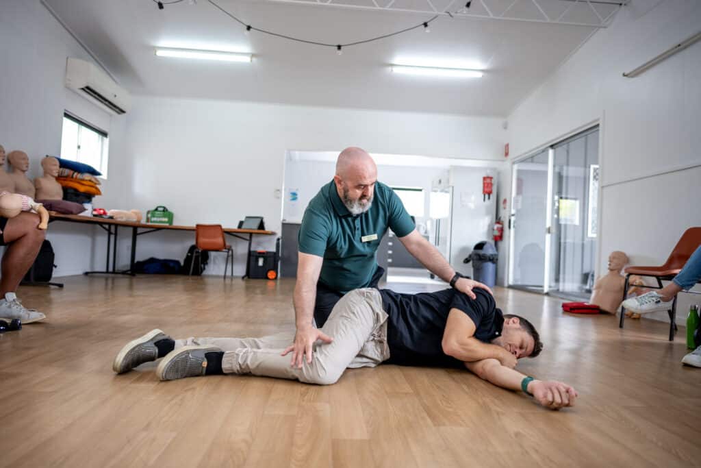 Step 3 of the recovery position: A first aid instructor adjusts the position of a person lying on the floor, completing the recovery position, with another person observing in the background.